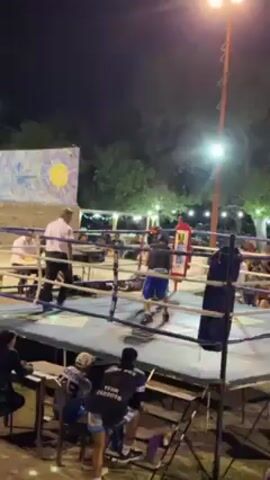The video captures a boxing match taking place at night. Two boxers, one in blue shorts and the other in grey shorts, are actively fighting in a boxing ring. The referee, dressed in a white shirt and dark pants, closely watches the fight, occasionally stepping in. Around the ring, spectators are visible, seated and standing, observing the match. A few people are sitting at a table adjacent to the ring. Trees and buildings are visible in the background, illuminated by streetlights. A large painting or mural depicting a sun is also present in the scene.