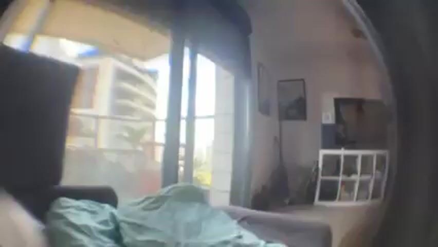 The video shows a fisheye lens view of an apartment interior. In the foreground, there's a brown couch with a light green blanket or sheet partially covering it. Behind the couch is a sliding glass door leading to a balcony with a partial view of a building across a distance. Inside the apartment, there's a shelving unit with various items, a television on top, and two framed pictures on the wall. A guitar and fan are situated near the wall. The lighting is generally bright, suggesting daylight. The overall impression is of a living space.