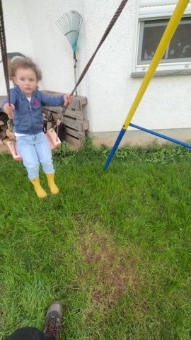 The video shows a small child on a swing in a grassy backyard. The child, with curly brown hair, is wearing a blue shirt, jeans, and yellow boots. The swing has a pink plastic seat and is suspended by brown ropes from a blue and yellow metal frame. The background features a white wall with a window, a rake leaning against the wall, and a wooden pallet. The ground is covered in green grass with some bare patches. The camera moves with the swing, keeping the child in focus.
