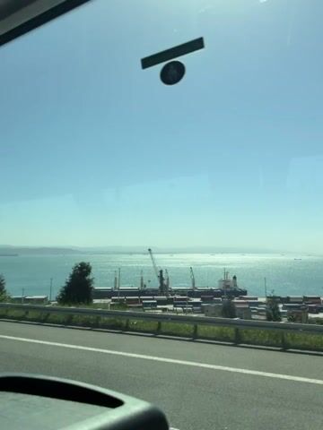 The video shows a bright, sunny daytime view from inside a moving vehicle, likely on a highway overlooking a large body of water. In the immediate foreground, the top of a vehicle's dashboard and reflections on the windshield are visible. A road with a metal guardrail separates the vehicle from the landscape.

Below the road, there's an active industrial port or harbor with numerous colorful shipping containers stacked, several large cargo ships docked, and multiple cranes for loading and unloading. Beyond the port, a vast expanse of water stretches towards the horizon, shimmering under the sunlight, with distant ships scattered across it. A green, tree-covered landmass is visible on the far side of the water.

The sky is clear and blue, with strong sun glare appearing at the top of the frame at various points. At the 0:01 mark, a large metal overhead gantry structure briefly passes over the road. Faintly visible text "COSCO" can be spotted on some containers. An unseen person is speaking in the background, mentioning "registration" and "Istanbul" in what appears to be a Slavic language.