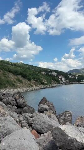 The video captures a static view of a seascape under a partly cloudy sky. The foreground is dominated by large, grey rocks, likely part of a breakwater or natural rocky shoreline. Beyond the rocks, a calm, blue body of water stretches towards the horizon. Along the coastline, a green, sloping hill is visible, dotted with white buildings, suggesting a coastal town or resort. The hill is covered in lush vegetation. In the distance, hazy mountains or hills are visible on the horizon. The sky is a bright blue, with fluffy white clouds scattered across it. The overall impression is one of a peaceful, natural coastal landscape on a clear day. There are no people or discernible actions in the video.