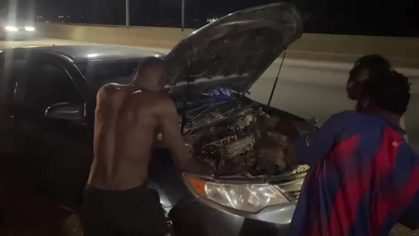 The video shows three individuals, two of whom are shirtless, gathered around the open hood of a dark-colored sedan. They appear to be working on the engine, suggesting a mechanical issue with the vehicle. The scene takes place at night, as indicated by the darkness and the presence of headlights from passing cars in the background. One of the individuals is wearing a blue and red striped shirt, possibly a sports jersey. The setting appears to be a roadside, possibly a highway or a bridge, given the elevation and the visible barriers. The overall mood is one of a roadside repair, with a sense of urgency or problem-solving. There is no visible text in the video. The location context provided is "Immigration Quarters, Nigeria."