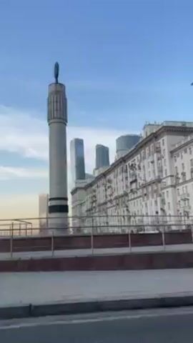 The video shows a drive-by view of a cityscape. In the foreground is a wide road with a sidewalk on the left side. There is a tall, cylindrical monument with a symbol at the top near the center of the frame. Behind it, there's a large, white building with many windows, suggesting an apartment complex. In the background, there are several modern skyscrapers, indicating a central business district. A black Mercedes drives past the camera. The sky is blue with some light clouds. A 30 speed limit sign is visible. The overall scene appears to be in a city like Moscow, based on the architectural styles and the presence of modern skyscrapers and the Soviet era monument.