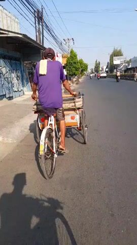 A person wearing a purple shirt, a dark cap, and plaid shorts pedals a three-wheeled bicycle with a passenger carriage on a paved road. A white towel is draped over the person's shoulders. The passenger carriage appears to be empty. The scene is outdoors on a sunny day, with buildings lining the left side of the road. A shadow of the person and bicycle is cast on the road. Other vehicles, including motorcycles, are visible in the distance on the road. Power lines are strung overhead.