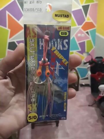 Here is a concise description of the video:

The video shows a hand holding a package of Mustad "Big Game" saltwater hooks. The package is blue and features images of fish and waves, along with the product name "Hoks Tripel." The hooks themselves are visible inside the package, featuring orange heads and pink and clear filaments attached to them. The package indicates they are size 5/0. The background is slightly blurred but appears to include a colorful wall and other miscellaneous objects.