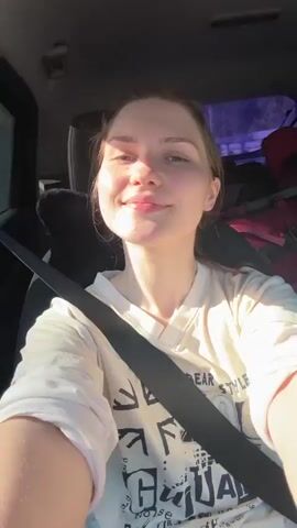 The video shows a young woman in the driver's seat of a car. She is wearing a white t-shirt and a seatbelt. She looks into the camera and smiles. The car appears to be in motion, and there are bags and other items in the backseat. The lighting suggests it's daytime.