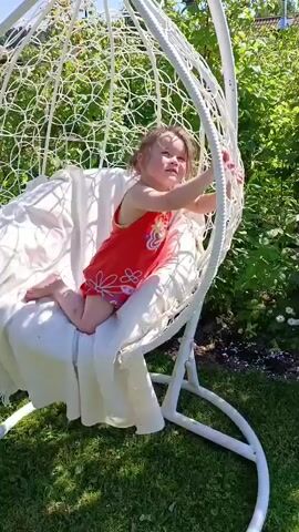 A young child wearing a red dress with a floral pattern sits on a white cushioned egg-shaped hanging chair. The child is on their hands and knees, facing towards the right side of the frame. The chair is outdoors, suspended from a white metal frame. Green grass and foliage surround the chair. A woman in a light green t-shirt and a beige skirt stands to the left of the chair, looking towards the child. She has bare feet. Two pairs of flip-flops are visible on the grass. The background shows more greenery, trees, and a bright blue sky with some clouds. The child is holding onto the edge of the chair with their right hand.