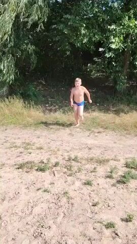 A young boy wearing blue swim trunks runs towards a body of water and jumps in, creating a large splash. He then submerges into the water. The scene is set in a natural environment with trees and grass surrounding the water.
