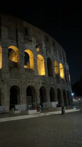 The video pans across the exterior of the Colosseum in Rome at night. The ancient structure is illuminated by warm yellow lights, highlighting its arches and weathered stone. Several people are visible sitting on steps in front of the monument, and a trash receptacle is present on the right side of the frame. The ground is paved with cobblestones and appears wet, reflecting the lights.