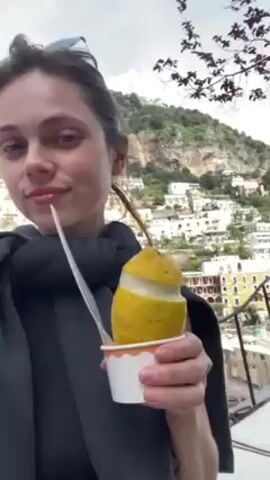 A woman is standing outside, holding a lemon filled with what appears to be gelato in a white paper cup, with a plastic spoon sticking out of it. She is smiling at the camera. She is wearing a black jacket or shawl. Sunglasses are perched on her head. In the background, there is a hillside town with closely packed buildings. The sky is overcast.