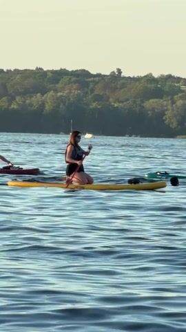 Two people are paddleboarding on a calm body of water with a tree-lined shore in the background. The person in the foreground is on a yellow paddleboard, kneeling and holding a paddle. They are wearing a dark life vest and dark shorts. To their right, another person is on a lighter-colored paddleboard, also kneeling and holding a paddle. They appear to be wearing a light-colored bathing suit. There is a red object on the paddleboard of the person in the foreground and a green object on the paddleboard of the person in the background. Several other paddleboards and a sailboat are visible in the distance. The sun is low in the sky, casting a warm light on the scene.