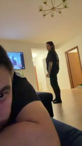 A man is lying down on a blue cushion with his head propped up. He is looking directly at the camera with one eye clearly visible. He has dark hair and a beard. Behind him, a woman with long, dark hair stands in the middle of the living room. She is wearing a black t-shirt and black leggings. She gestures with her right hand towards her wrist, as if checking the time. The living room has wooden floors, a television on the wall, and a light fixture on the ceiling. There are doorways leading to other rooms in the background. The woman places her hands on her hips and stands up straight.