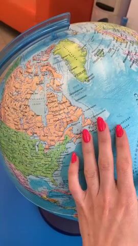 The video shows a close-up of a hand with bright red fingernails resting on a spinning globe. The globe displays continents and oceans. The hand appears to be touching the surface of the globe, with fingers spread across the landmasses and water. The background is blurred but suggests an indoor setting with hints of orange and blue. The lighting is bright, and the focus is on the globe and the hand.