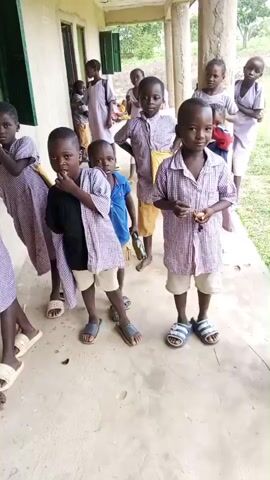 A group of school-aged children, primarily young boys and girls, are standing in a line along the veranda of a building. The children are wearing school uniforms, consisting of checkered shirts and shorts or skirts. Some of the children appear to be looking at the camera, while others are engaged in conversation or looking away. The environment appears to be an outdoor setting, with a building wall and pillars visible, as well as some greenery in the background. The lighting suggests it is daytime. The children are standing on a concrete floor.
