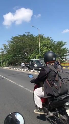 The video is a first-person perspective of someone riding on the back of a motorcycle on a paved road. The rider in front is wearing a black helmet, a black jacket, and a backpack. The motorcycle is black with a white license plate. To the right of the road, there is a sidewalk with some people on motorcycles and what appears to be a truck. The background consists of trees and a blue sky with white clouds. The sun is shining, casting shadows. The road has white lines marking lanes and a yellow and black striped barrier. The overall environment appears to be a street in a tropical or warm climate.