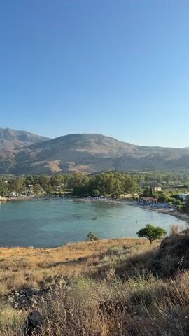A wide shot shows a bay with calm, turquoise water. Mountains rise in the background, with a small town nestled at the base of the hills. A sandy beach with scattered umbrellas and sunbathers is visible along the bay. In the foreground, dry, golden grass covers a hillside. On a small rocky outcrop in the bay, a tiny white church with a red tile roof stands out. The sky is clear and blue. The video pans slightly to the left, revealing more of the coastline and the distant mountains.