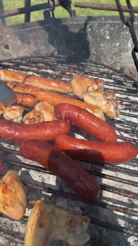 A man barbecues sausages and chicken wings on a circular grill suspended by chains over a stone fire pit. He is kneeling on the grass in a backyard, wearing a two-toned t-shirt and black shorts. He uses silver tongs to flip the food, while smiling at the camera. The grill contains an assortment of sausages, including some reddish-brown ones and some lighter-colored ones, as well as chicken wings with crispy, browned skin. A silver pot sits beside the grill, and a green lawn and modern house are visible in the background under a clear blue sky.