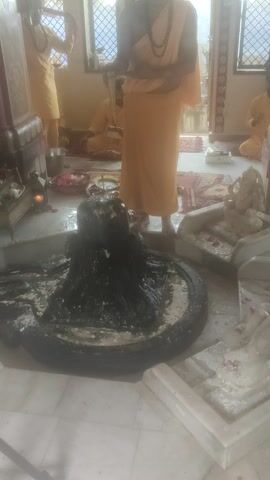 The video shows a Hindu temple interior with several individuals in orange robes. In the foreground, a person is standing and appears to be performing a ritual at a Shiva Lingam, which is covered in a dark substance and surrounded by offerings. Another person is also standing near the Lingam. In the background, other individuals in similar attire are either kneeling or standing, also appearing to participate in religious activities. The environment is dimly lit, with windows providing natural light. The walls are decorated with intricate patterns. The floor is tiled. There are various religious items and decorations visible. The camera angle is skewed, making it difficult to determine the exact orientation of the room.