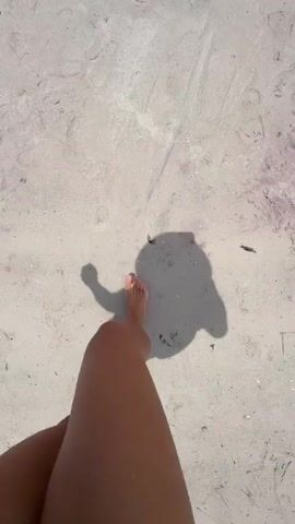 This video shows a person walking on a sandy beach. The camera is positioned above and behind the person, looking down at their legs and feet as they walk. The sunlight casts a prominent shadow on the sand, which also moves with each step. The sand appears light in color and has some small debris scattered on it, suggesting a natural beach environment. The person's legs are tanned, and they appear to be wearing shorts or a swimsuit, indicating a warm-weather setting, likely during the day given the strong shadow. The overall mood is relaxed and leisurely, consistent with a beach stroll. The scene is likely in North Miami, United States of America. There is no visible text in the video.