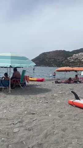 The video shows a pebble beach with people relaxing under umbrellas and engaging in water activities. In the foreground, there is a gray pebble beach with several small rocks scattered across it. To the left, an umbrella with blue and white stripes provides shade for a few people sitting at a table. A red and yellow kayak is also visible on the beach near the umbrella.

In the middle ground, the water is calm and clear, with several individuals enjoying paddleboarding. One person is standing on a paddleboard in the distance, while another is seated on a paddleboard closer to the shore. A small orange and white paddleboard with a black fin is positioned on the beach in the right foreground.

Further back, a gentle slope rises from the water, covered in green vegetation and dotted with white houses. A cylindrical tower is visible on the crest of the hill. The sky is overcast, with some clouds. The overall scene depicts a peaceful day at a coastal resort.