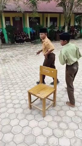 The video shows two young boys in school uniforms, each wearing a black cap, engaged in a game. One boy in a tan uniform is on the left, and the other in a light green uniform is on the right. A single wooden chair is placed in the foreground between them. The boys are positioned on a patterned stone surface, possibly a schoolyard, with a building in the background. In the background, a group of people are seated, appearing to be an audience. The boy on the left is bent over and appears to be moving his legs as if dancing or performing a ritualistic movement. The boy on the right is standing and watching the boy on the left. The scene suggests an outdoor school event or activity.