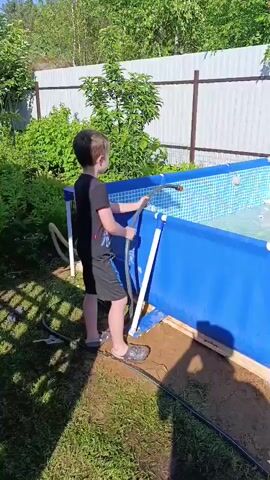 A boy stands next to a blue frame pool and fills it with water from a hose. He is wearing a black t-shirt and black shorts. The pool is filled with water, and there are some toys floating in it. The pool is in a backyard, with a fence and trees in the background. The weather is sunny.