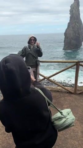 A person in a black hoodie takes a photo with a smartphone of another person wearing a green jacket, beige pants, a white hood, and sunglasses, who is posing in front of the ocean with a large rock formation in the background. The person taking the photo has a light green purse hanging over their shoulder. They are both standing on a dirt surface with a wooden fence separating them from the ocean's edge. The weather appears cloudy and overcast.