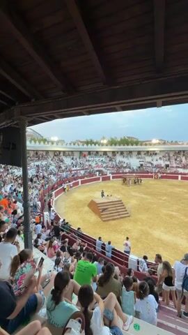 This video captures a wide-angle view from the stands of a bullring, looking down into the sandy arena where several individuals are gathered. The stands are filled with spectators, many of whom are seated and facing the arena. The architecture of the bullring appears traditional, with wooden beams supporting the roof structure visible in the foreground. The lighting suggests it is late afternoon or early evening, with a slightly overcast sky. The overall scene depicts a public event taking place within a specialized sporting venue.