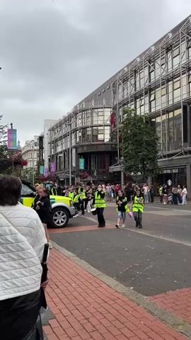 The video shows a street scene with several people walking and standing. In the foreground, a person wearing a white puffer jacket is seen from behind. To the left, part of a yellow and white vehicle, possibly an ambulance or service vehicle, is visible. Several individuals wearing bright yellow high-visibility vests are walking in the street. One is carrying a bucket. Two young women in similar vests are walking together, each holding a water bottle. Further in the background, a man in a dark shirt and jeans is walking, carrying a bag over his shoulder. There are buildings with modern architecture lining the street, one of which has large windows. A colorful mural is visible on a wall in the background. There are also trees and hanging flower baskets. The sky is overcast. The overall impression is of a public event or gathering taking place on a city street.