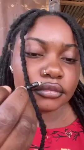 A close-up shot shows a Black woman with dark dreadlocks wearing a red and black patterned shirt. She is applying black lip liner with a black pencil. The background is slightly out of focus and appears to be a simple outdoor setting. The woman has a nose ring and small pearl earrings.  Her expression is focused as she applies the lip liner.