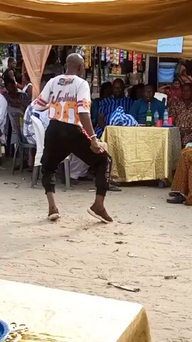 Under a canopy at an outdoor event, a bald, dark-skinned man in a white t-shirt with the words "Worldwide 80" printed on the back, black pants with red stripes down the side, and brown shoes, is dancing. He is facing away from the camera, towards a group of people sitting at tables.  The ground is sandy, and there are various objects scattered around, including a blue plastic plate on a table in the foreground. In the background, people are seated at tables, and there's a small shop with various items on display. Two young boys in matching red shirts walk past the dancing man carrying a black plastic bag.