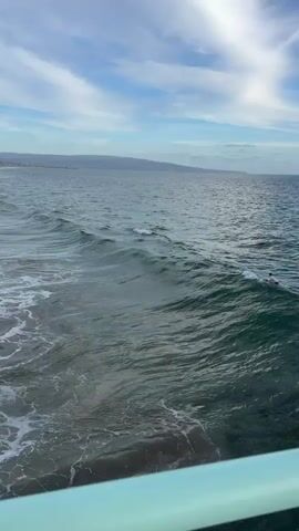 The video begins with a view of the ocean from a pier. A surfer is visible in the distance to the right. As the video progresses, a wave approaches and begins to break, creating white foam and churned water. The camera pans slightly to the left, revealing the beach in the background with people walking and sunbathing. The sky is partly cloudy. The video ends as the wave continues to break.