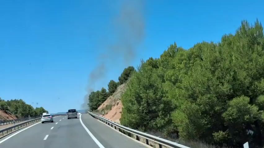 The video shows a bright, sunny day on a highway in Ariza, Spain. The camera is positioned inside a moving vehicle, looking forward. The highway is asphalt with white lane markings. On the right side of the road, there is a steep, reddish-brown embankment covered in green pine trees and dry brush. A metal guardrail runs along the edge of the road. Several cars are visible on the highway, including a white sedan and a darker SUV, as well as a large truck in the distance.

The most notable feature of the scene is a large plume of dark grey smoke rising from the hillside on the right side of the highway, partially obscured by trees. The smoke suggests a fire is occurring off-camera, likely in the vegetation on the slope. The sky is a clear, vibrant blue, indicating fair weather. The time of day appears to be late morning or early afternoon due to the bright sunlight and the angle of the shadows. There is no visible text in the video. The overall mood is one of travel on a clear day, with the unsettling presence of the smoke hinting at an environmental concern.