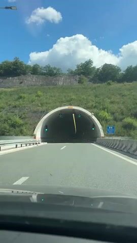 The media shows a view from inside a car driving through a tunnel. The tunnel is well-lit with rows of rectangular fluorescent lights on the ceiling. The walls are white with a red stripe running horizontally. There are also green traffic lights visible at the entrance of the tunnel and red lights spaced along the tunnel. The road is asphalt with white lane markings. On the right side of the tunnel entrance, there is a blue road sign with white text.

The scene appears to be during the day, with bright sunlight visible at the tunnel entrance. The sky is blue with some white clouds. The area surrounding the tunnel entrance is covered in green vegetation and trees. The overall impression is one of modern infrastructure facilitating travel.

Visible text on the sign reads: "Бањаречка Бања 733 m". This indicates that the tunnel is on the way to Banjačka Banja, and it is 733 meters long. Given the location context of "Градска општина Нишка Бања, Serbia", this suggests the tunnel is located near Niška Banja in Serbia, and likely leads towards the Banjačka Banja area.

The activity taking place is vehicular travel through a tunnel. There are no people visible outside of the car. The emotion evoked is likely one of passage and progress, typical of a journey through a tunnel.