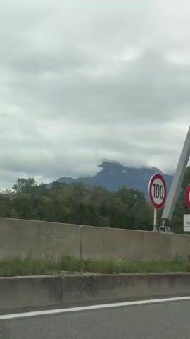 The video shows a view from a vehicle traveling on a highway. The highway has a concrete barrier separating it from the opposing lanes or a shoulder. Beyond the barrier, there is vegetation and trees, and in the distance, mountains can be seen. The sky is overcast with clouds. Several vehicles are visible on the highway, including a white van with writing on the side, a truck, and a few cars. There is also an overhead pedestrian bridge crossing the highway. The camera appears to be looking forward and slightly to the right, capturing the passing scenery. The overall impression is of a journey through a landscape with mountainous terrain under a cloudy sky.
