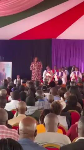 A medium shot shows a woman speaking at a podium to a crowd seated in rows of chairs. The woman is wearing a long-sleeved floral dress. Behind her, seated on a raised platform, are approximately ten people in formal wear, including several men in purple robes. The crowd in front of the speaker is diverse in age and dress. Overhead is a canopy with red, white, and green stripes. To the left of the speaker is a poster on an easel. A black curtain and purple draperies serve as a backdrop. The speaker gestures with her hands as she addresses the crowd. The scene appears to be an event or gathering, possibly a church service or memorial.