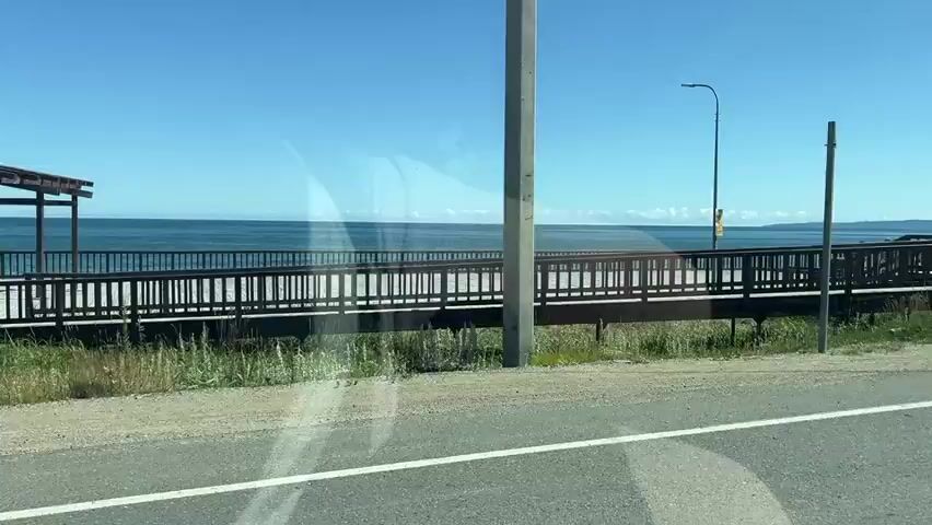 The video shows a view from a moving vehicle, looking towards a scenic coastline on a clear, sunny day. In the foreground, a paved road with a white line is visible. Adjacent to the road is a grassy area and a wooden boardwalk running parallel to the coastline. The boardwalk features wooden benches and covered seating areas. The ocean stretches out to the horizon, with a clear blue sky above. In the distance, landmasses can be seen on the horizon. There are a few people visible on the boardwalk, some walking and others sitting. A street light and a road sign are also present. The overall atmosphere is peaceful and serene, suggesting a leisurely drive along a coastal route.