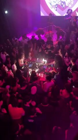 A crowded nightclub has a DJ playing music on multiple turntables behind a safety barrier. The dancefloor is packed with people who are dancing and holding drinks. A projector screen behind the DJ shows an animated graphic in pink, white and purple. The club is illuminated with bright purple and red lights. Some people are looking at their phones.