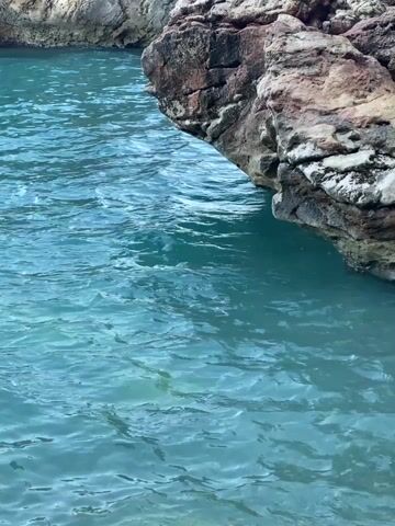 The video shows a close-up view of clear blue water lapping against a rocky shoreline. The water is calm and translucent, revealing the sandy bottom beneath. The rocks are rough and irregular, with shades of brown, gray, and white. The scene is tranquil and appears to be a natural coastal environment.