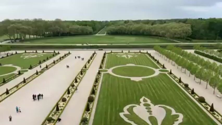 Here is a concise description of the video:

The video presents an aerial view of a meticulously landscaped garden. The garden features geometric patterns created with grass and gravel pathways. People are walking along the pathways. Beyond the formal garden, there is a view of a large open green space and treeline. The sky is overcast.