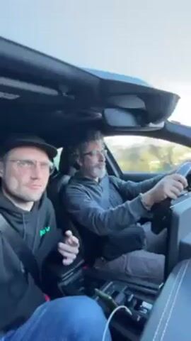 The video shows the interior of a car with two men driving. The driver is middle-aged, has graying curly hair and is wearing glasses, a gray long-sleeved shirt, and pants. The passenger is younger with blond hair and glasses. He is wearing a black hoodie and a baseball cap. The car has a sunroof and black leather interior. There is loud techno music playing in the background. The car is in motion.