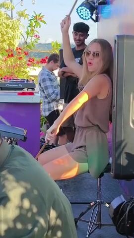 A young woman with long brown hair sits on a silver stool holding drumsticks, ready to play. She's wearing a light brown romper and sunglasses. Behind her, a smiling man watches with arms crossed. Another man in a plaid shirt stands near a purple DJ booth with a Pioneer DJ mixer. A large black speaker stands to the right. The setting appears to be an outdoor event, possibly on a rooftop, with plants and string lights visible.