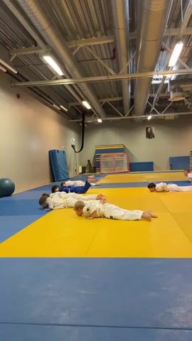 The video shows a group of children practicing judo in an indoor training facility. The children are wearing white judogi (judo uniforms) with yellow belts. They are lying on a blue and yellow mat, with some of them in a prone position and others in a push-up position. A man in a blue judogi is kneeling on the mat, facing the children, appearing to instruct them. The room has a high ceiling with exposed pipes and lighting fixtures. In the background, there are large blue mats leaning against a wall, a blue exercise ball, and what appears to be a hockey goal. The overall scene suggests a judo class for young children, likely during daylight hours given the interior lighting. There is no visible text in the video.