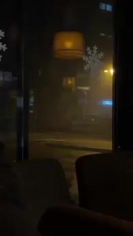 From the perspective of someone sitting at a table inside a cafe, a nighttime view of a city street on a foggy night is visible through a large window. Snowflake decals and an outline of Santa Claus are on the glass. A street light glows in the fog. Several cars pass on the street.