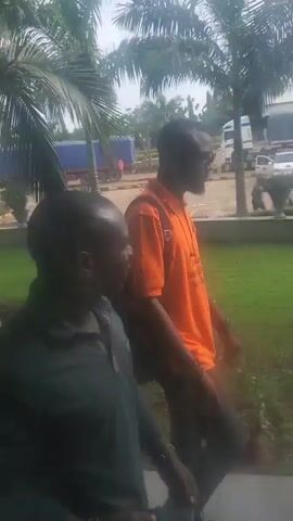 The video shows a group of men walking. The camera is moving, and the focus shifts between the men and the surrounding environment. In the foreground, a person in a dark shirt is partially visible. Further back, another person in an orange shirt with a black backpack walks past. Beyond them, a white van is parked, and several palm trees are visible. In the background, a building with a yellow and white facade is present, along with other vehicles. Later in the video, a man in a light blue shirt and dark pants walks on a paved path, carrying a dark bag. The environment appears to be an outdoor setting with greenery, trees, and paved areas.