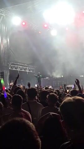 A concert crowd is visible in the foreground, with many people holding up glowing sticks in various colors. The stage is in the background, with bright red and white lights illuminating it. A performer is on stage, singing into a microphone. A large screen behind the performer shows a black and white image of the performer as well. Smoke or fog is visible on the stage, adding to the atmospheric lighting. The overall scene suggests an energetic and lively musical performance.