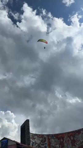 A paraglider is captured in mid-flight against a backdrop of dramatic, cloudy skies. The paraglider, a person strapped into a harness, is suspended beneath a colorful wing, predominantly yellow and red. The wing is angled upwards, suggesting the paraglider is either ascending or gliding at an upward trajectory. The sky is filled with thick, gray clouds, some appearing wispy and others more solid, creating a sense of depth and an overcast atmosphere. The lower portion of the video shows the tops of some structures, including what appears to be a stone tower on the left and a brightly colored mural or painted wall on the right, adorned with intricate figures and patterns. The overall scene conveys a sense of adventure and the vastness of the sky.
