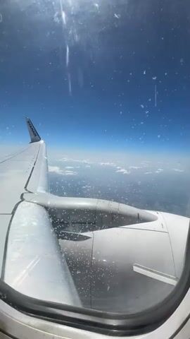 A video shows a view from an airplane window. The wing of the plane is visible on the left side of the frame, with the Ryanair logo on the wingtip. Below the wing, the engine is visible. The sky is a bright blue with scattered clouds, and the ground can be seen through the clouds as a hazy green and brown landscape. There are water droplets or condensation on the inside of the airplane window. The video captures a brief moment of flight, with the plane appearing to be in cruise altitude.
