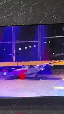 The video captures a robot combat event, likely a match between two robotic competitors. In the foreground, the arena floor is visible, illuminated by blue and purple stage lighting. A large, metallic robot, emblazoned with the word "BRONCO" in red lettering, is being flipped upside down and tossed by an unseen force, its underside and wheels clearly visible. To the left, a smaller, dark-colored robot with a red accent on its front is on the arena floor, appearing to be stationary or having just been impacted. The background features a high-tech arena with glass walls and audience seating, also bathed in dramatic lighting. The overall scene is dynamic and action-oriented, suggesting a moment of intense physical engagement between the robotic combatants.