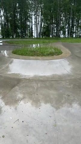 The video opens with a shot of a wet, empty skatepark. The concrete surfaces are glistening with water, indicating recent rain. The park features smooth, curved ramps and bowls, surrounded by lush green grass and a dense line of tall birch trees. In the distance, through the trees, a body of water is visible, and the sky appears overcast, suggesting a cloudy or rainy day.

As the video progresses, two individuals enter the frame, walking through the skatepark. They are dressed in casual, dark clothing. One person carries what appears to be a skateboard or a similar piece of equipment. The other person walks beside them, and they seem to be engaged in conversation or simply moving through the space together. The overall mood conveyed is subdued, possibly due to the weather and the quiet atmosphere of the deserted park. The presence of the water and the surrounding natural elements create a somewhat serene, albeit damp, environment. No visible text is present in the video.